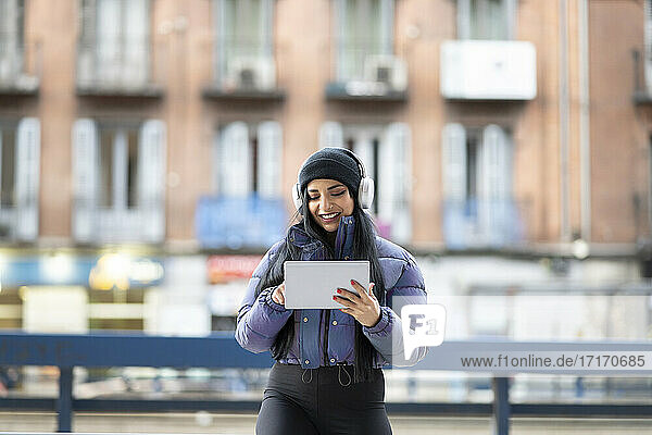 Smiling young woman using digital tablet while listening music against buildings in city