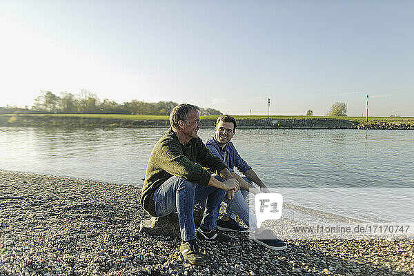 Father and son spending leisure time sitting at riverbank against clear sky