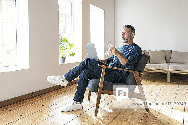 Nachdenklicher Geschäftsmann  der eine Brille hält  während er zu Hause am Laptop sitzt