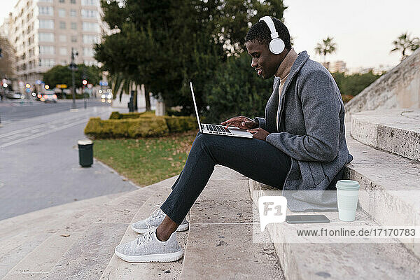 Junger Mann mit Kopfhörern  der einen Laptop benutzt  während er auf einer Treppe sitzt