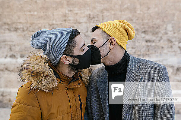 Homosexuelles Paar mit Gesichtsschutz  das sich küsst  während es an der Wand steht