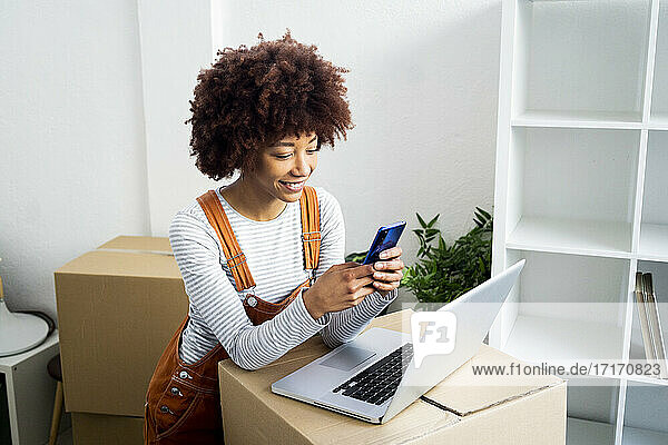 Afro-Frau benutzt ihr Smartphone  während sie sich in ihrer neuen Wohnung auf einen Karton stützt