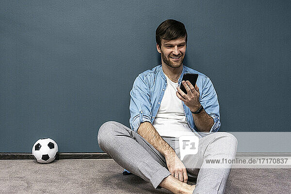 Lächelnder Mann beobachtet Fußball auf dem Handy  während er an einer grauen Wand sitzt