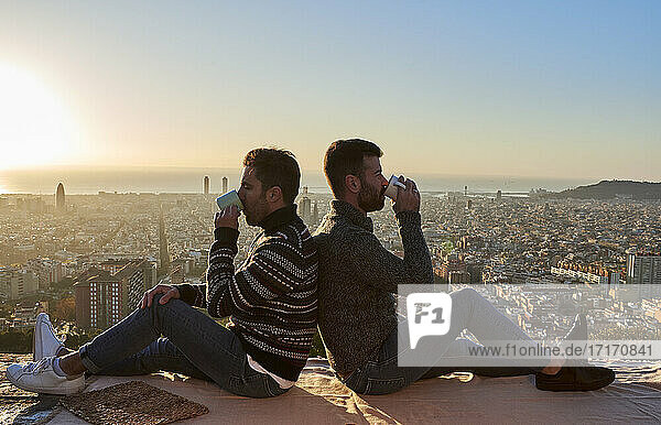 Gay couple drinking coffee while sitting back to back on observation point at Bunkers del Carmel  Barcelona  Spain