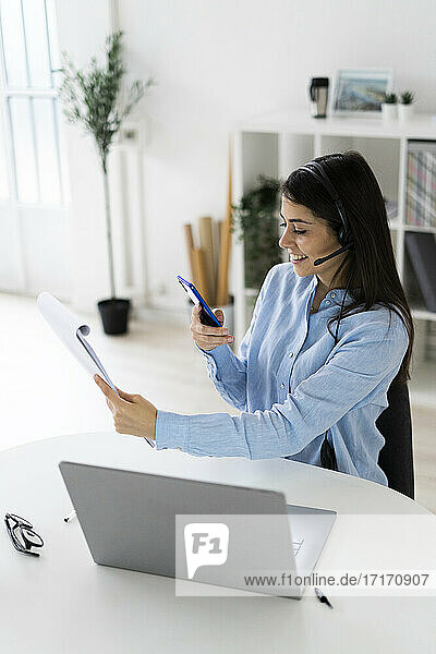 Geschäftsfrau mit Headset  die einen Notizblock mit ihrem Mobiltelefon fotografiert  während sie am Schreibtisch im Büro sitzt