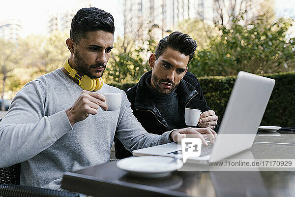 Male friends looking at laptop while having coffee sitting at sidewalk cafe at street