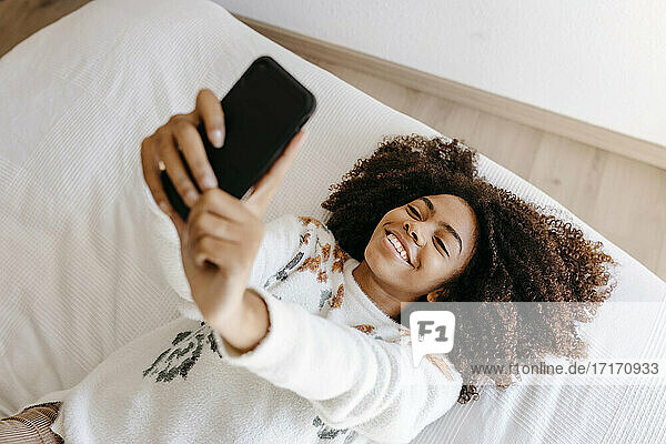 Lächelnde Frau  die ein Mobiltelefon benutzt  während sie zu Hause auf dem Bett liegt