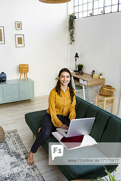 Smiling woman working on laptop while sitting on sofa at home