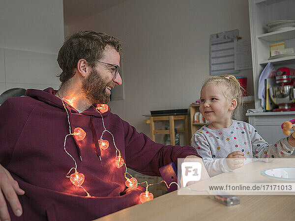 Smiling father with string light around neck while looking at daughter eating in kitchen