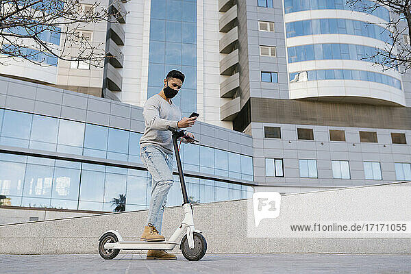 Young man with electric scooter using smart phone while standing against buildings in city during COVID-19
