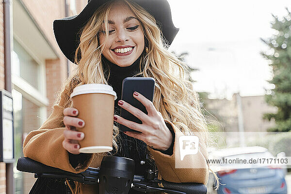 Happy young woman using smart phone while holding reusable coffee cup