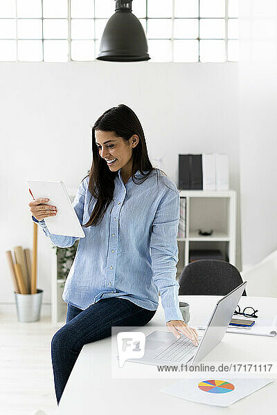 Geschäftsfrau  die an einer Telefonkonferenz auf einem digitalen Tablet teilnimmt  während sie im Büro am Laptop sitzt