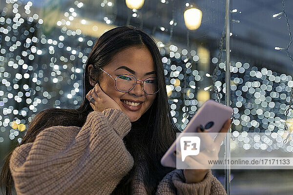 Smiling young woman with hand on chin using smart phone against illuminated glass wall