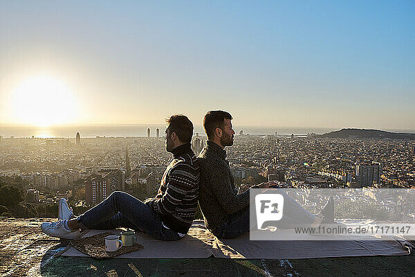 Gay couple sitting back to back on observation point during sunrise  Bunkers del Carmel  Barcelona  Spain