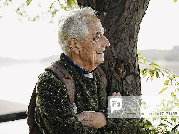 Smiling man with backpack looking away while leaning on tree