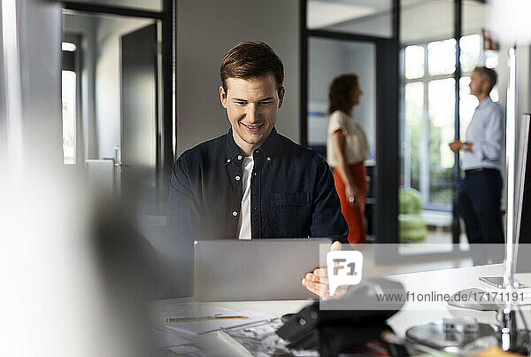 Lächelnder Geschäftsmann  der ein digitales Tablet benutzt  während er mit Kollegen im Hintergrund in einem Großraumbüro sitzt