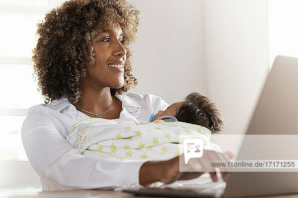 Lächelnde Frau, die am Laptop arbeitet, während sie mit ihrem Baby im Home Office sitzt