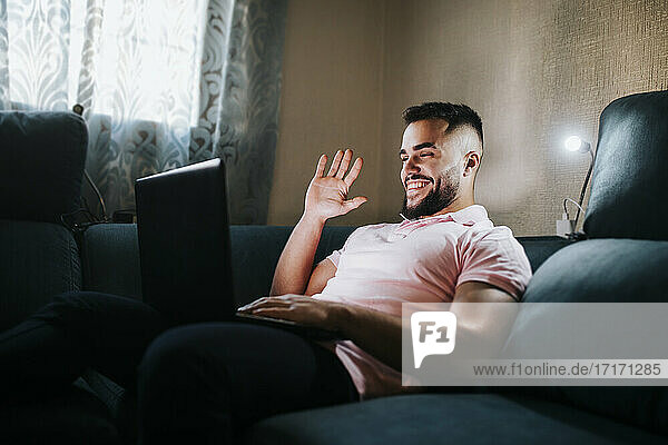 Young businessman waving hand on video call through laptop while sitting on couch in living room
