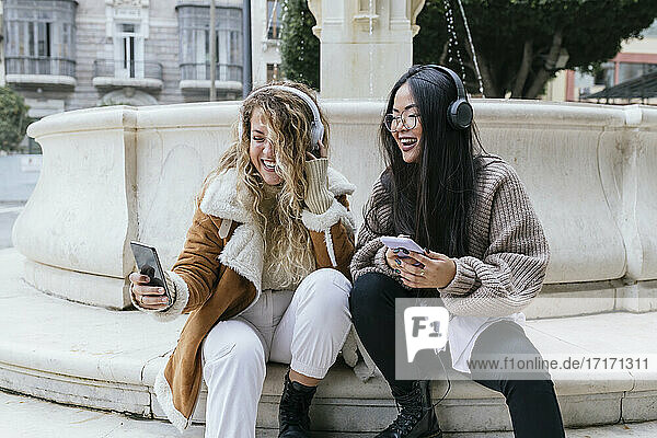 Laughing female friends taking selfie wearing headphones while sitting against fountain