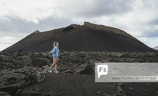 Weiblicher Tourist schaut weg  während er mit ausgestreckten Armen auf dem Berg am Vulkan El Cuervo steht  Lanzarote  Spanien