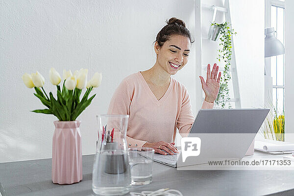 Young woman waving hand to video call on laptop while sitting at home office