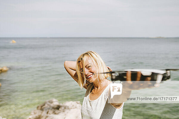 Lächelnde Frau  die eine Drohne hält  während sie am Meer steht