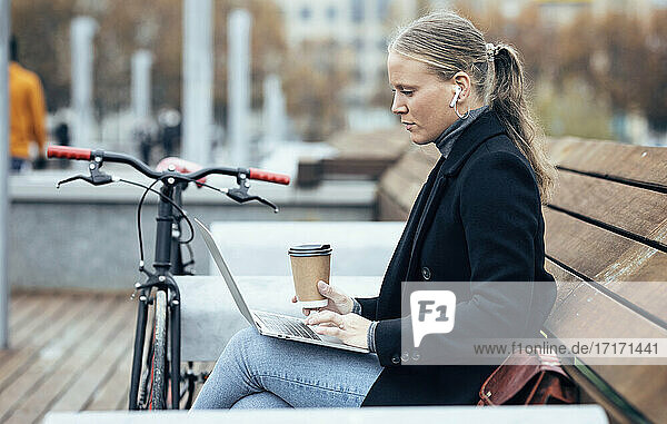 Woman in trench coat using laptop while sitting on bench by bicycle