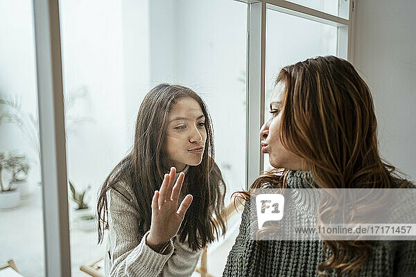Mother and daughter puckering while looking at each other through glass window at home