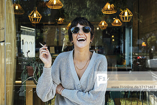 Young woman wearing sunglasses smoking cigarette while standing against restaurant window