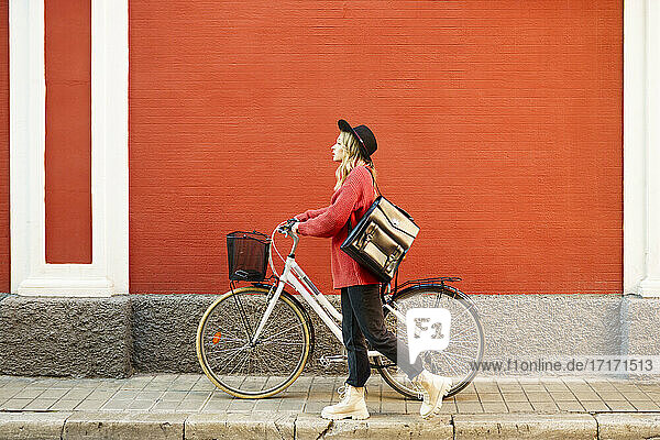 Young woman wearing hat pushing bicycle in front of red wall