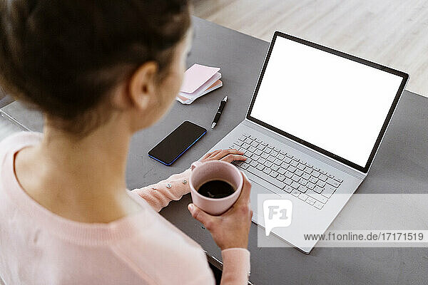 Young woman holding coffee cup while working on laptop at home