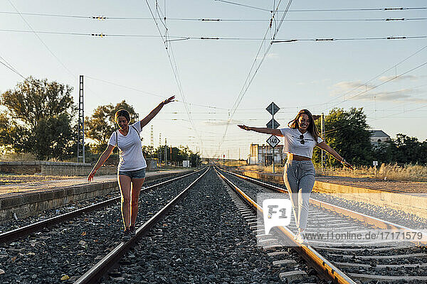 Unbekümmerte Freundinnen mit ausgestreckten Armen  die auf einer Eisenbahnstrecke gegen den Himmel bei Sonnenuntergang laufen
