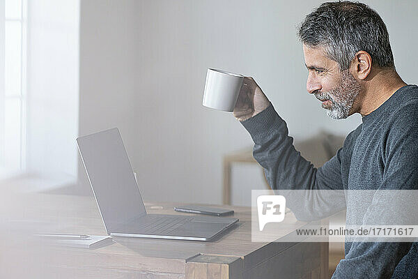 Geschäftsmann mit Kaffeetasse im Gespräch über Videoanruf durch Laptop  während er zu Hause sitzt