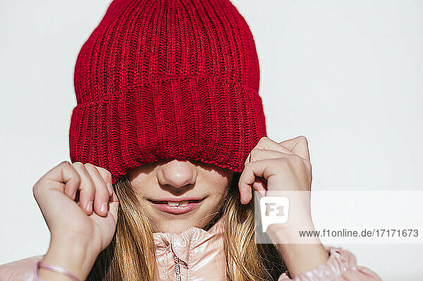 Playful girl covering eyes through knit hat against wall