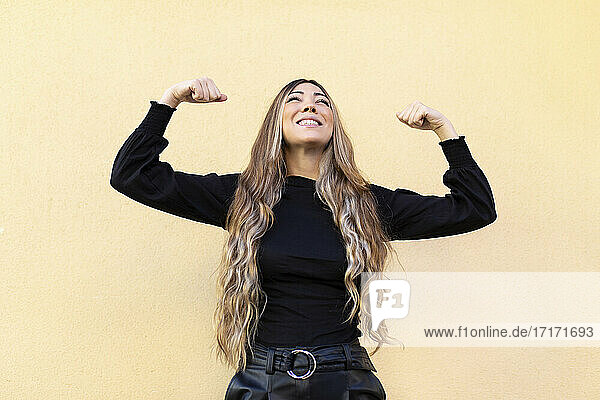 Junge Frau mit langen Haaren  die ihre Muskeln anspannt  während sie vor einem beigen Hintergrund nach oben schaut