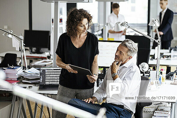 Mature business people using digital tablet while working with colleagues in background at open plan office