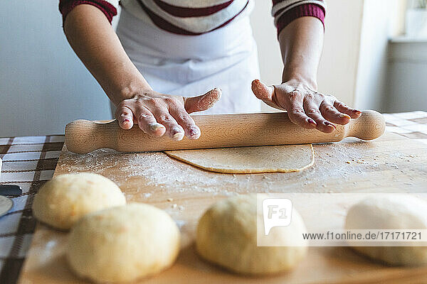 Woman flattening dough with rolling pin on cutting board to make croissants in kitchen