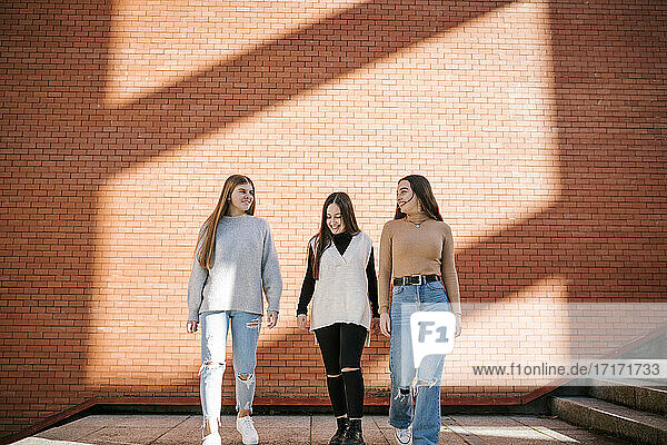 Lächelnde Freundinnen  die auf dem Fußweg gegen eine Backsteinmauer in der Stadt laufen