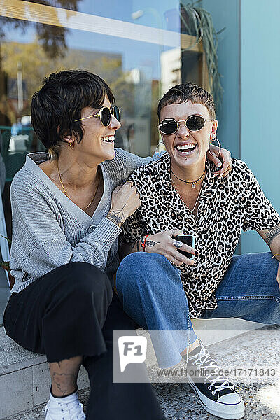 Smiling woman sitting with arm around on happy friend while sitting on footpath