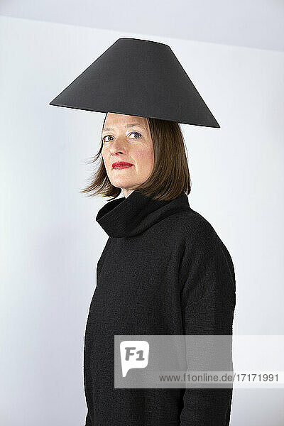 Mature woman with black lamp shade on head against white wall