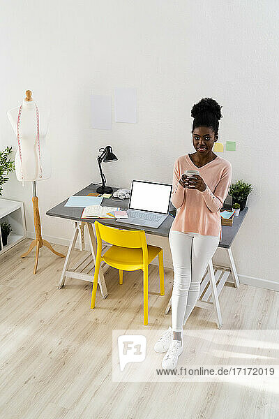 Lächelnde Modedesignerin trinkt Kaffee im Stehen im Büro zu Hause