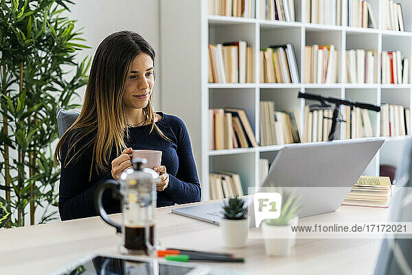 Weibliche Freiberuflerin schaut auf ihren Laptop und hält eine Kaffeetasse in der Hand