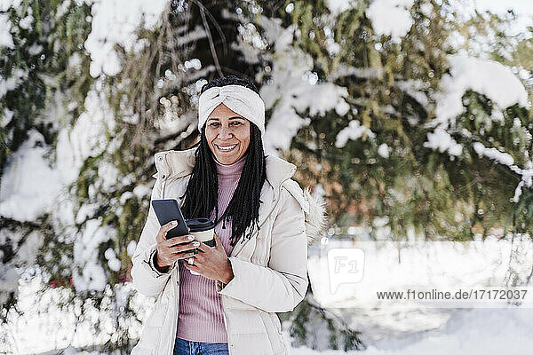 Smiling woman with smart phone and coffee cup in park during winter