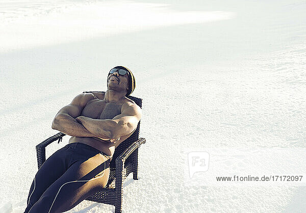 Mid adult sportsman wearing sunglasses resting while sitting with arms crossed on chair in snow