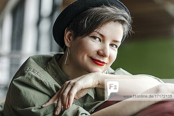 Lächelnde Frau mit Hut sitzt mit der Hand am Kinn auf einem Sofa im Studio