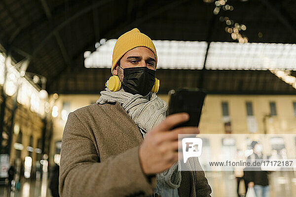 Mann mit Schutzmaske und Strickmütze  der am Bahnhof steht und ein Mobiltelefon benutzt