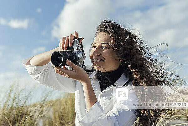 Smiling young woman photographing through vintage camera at beach against cloudy sky