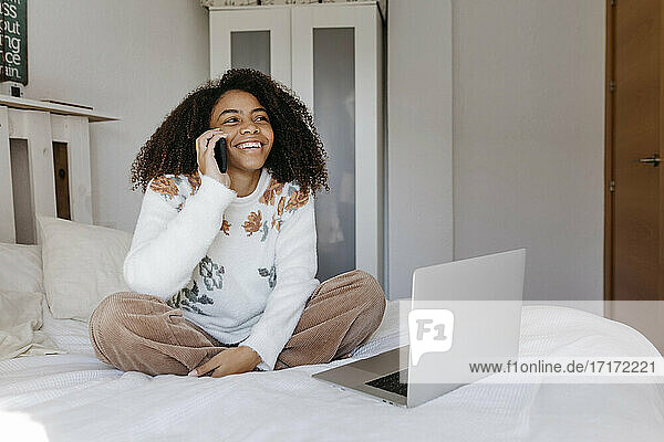 Lächelnde Frau mit Laptop  die zu Hause sitzt und mit ihrem Handy telefoniert