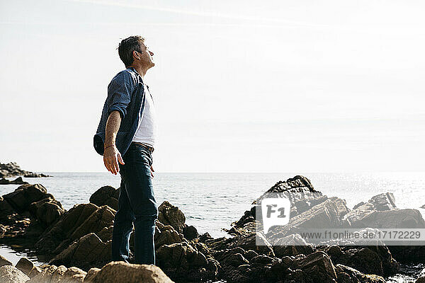Genießen Sie reifen Mann stehend inmitten von Felsen am Meer während sonnigen Tag