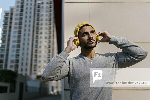 Thoughtful man listening music on headphones while looking away against wall during sunny day